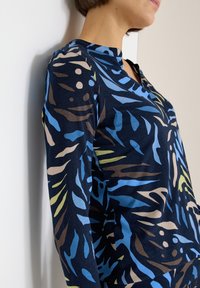 Bluzka z długim rękawem w kolorze granatowym z żywym, abstrakcyjnym wzorem w odcieniach niebieskiego, zielonego i brązowego. Miękka tkanina, okrągły dekolt i dopasowany krój.
