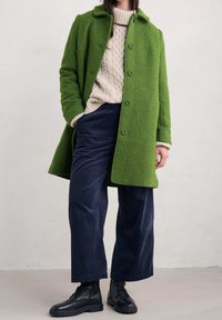 Zielony, teksturowany wełniany płaszcz z kołnierzem, z zapinanym przodem i bocznymi kieszeniami, nałożony na kremowy sweter w warkocze i granatowe spodnie z kordurowej tkaniny. Czarne buty sznurowane.