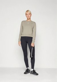 Sweatshirt cinzento com logótipo Nike, leggings pretas e sapatilhas desportivas pretas. A modelo segura uma garrafa de água preta, em pé sobre um fundo neutro.