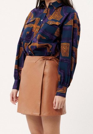 Femme portant une chemise bleu marine avec des motifs géométriques orange et une mini-jupe en cuir beige avec un détail de ceinture nouée.