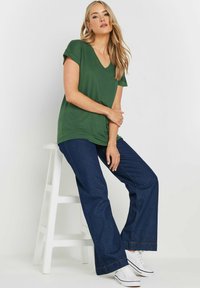 Grön v-ringad t-shirt med uppvikta ärmar i kombination med vida mörkblå denimjeans och vita sneakers, sittande på en vit pall.