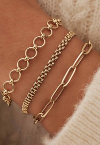 Trois bracelets en or avec des designs différents : une chaîne maillée, une chaîne de perles carrées et des maillons ovales allongés, tous présentés sur un poignet.