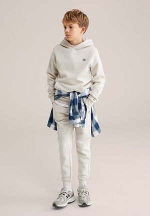 Svetlá mikina s malým logom, spojená s nohavicami so zúženým strihom. Modrá károvaná košeľa viazaná okolo pása. Neutrálny typ tenisky dopĺňa vzhľad.