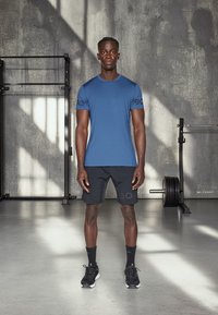 Modra športna majica in črne kratke hlače, z logotipom, v kombinaciji s črnimi športnimi copati in nogavicami. Gladko blago, minimalističen dizajn, okolje v telovadnici.