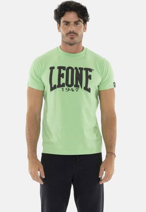Homme aux cheveux foncés portant un t-shirt vert clair à manches courtes avec "LEONE 1947" en lettres noires et un pantalon noir, debout devant un fond blanc.
