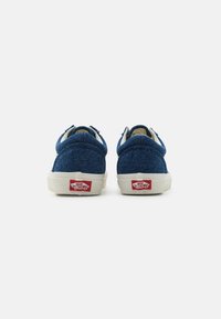 Blue denim sneakers with white rubber soles, rounded toe, and contrasting red logo at the heel. Textured fabric and simple design.