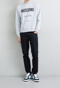 Šedá mikina s potiskem "MOSCHINO COUTURE! Milano", doplněná černými kalhotami a vícebarevnými teniskami, stojící na světlém pozadí.