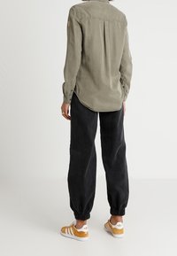 Olivezielona koszula z długim rękawem z plisą z tyłu, zestawiona z czarnymi, zwężanymi spodniami i białymi trampkami z żółtymi akcentami.