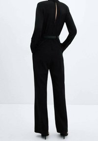 Combinaison noire avec une taille cintrée, des manches longues et une découpe dans le dos. Fabriquée en tissu lisse, elle présente un design à jambes larges et un minimum de détails.