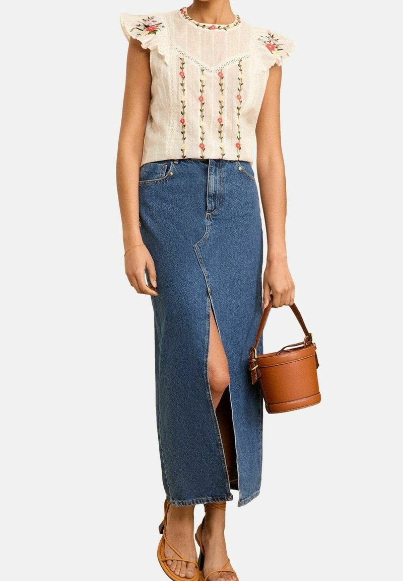 Femme portant un chemisier blanc brodé de fleurs, une jupe en denim bleu taille haute fendue, des sandales marron clair, tenant un sac seau en cuir marron.