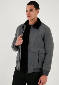 Sivá bomber bunda s čiernym golierom, prednou vreckom, ribbovanými manžetami a zipsom. Vyrobená z textúrovanej látky s hladkým povrchom.