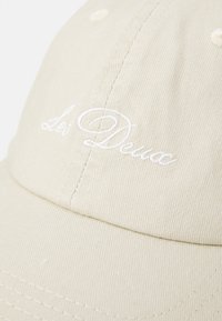 Beige baseballcaps med hvit brodert tekst "Les Deux" på frontpanelet, som viser tekstur i stoffet og detaljene i sømmen.