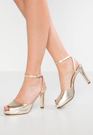 Złote szpilki peep-toe o gładkim metalicznym wykończeniu, z paskiem wokół kostki z klamrą i smukłym obcasem typu stiletto. Gładka tekstura na całej powierzchni.