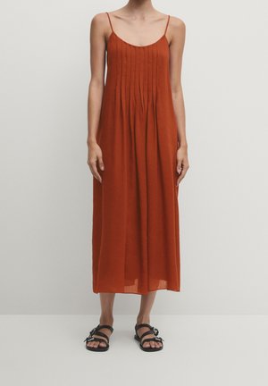 Woman wearing a rust-colored pleated midi dress with thin straps and black flat sandals, standing against a plain light background.