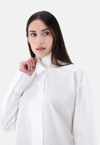 Camicia bianca a maniche lunghe realizzata in tessuto morbido con colletto alto, dotata di chiusure con bottoni e vestibilità leggermente oversize.