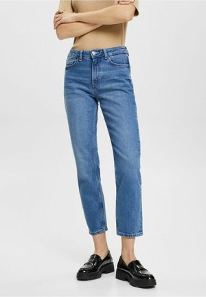 Osoba w niebieskich, krótkich, prostych dżinsach, beżowej bluzce z krótkim rękawem i czarnych, błyszczących mokasynach, z założonymi na klatce piersiowej ramionami.