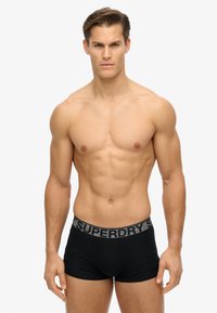 Bañadores de boxeo negros con una cinturilla gris que presenta el logo "SUPERDRY". Textura suave, forma ajustada y diseño minimalista.