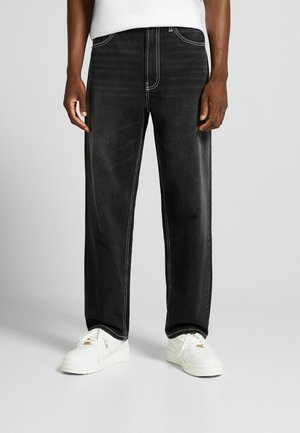 Personne portant un jean noir à jambes droites avec des coutures blanches et des baskets blanches, debout devant un fond gris.