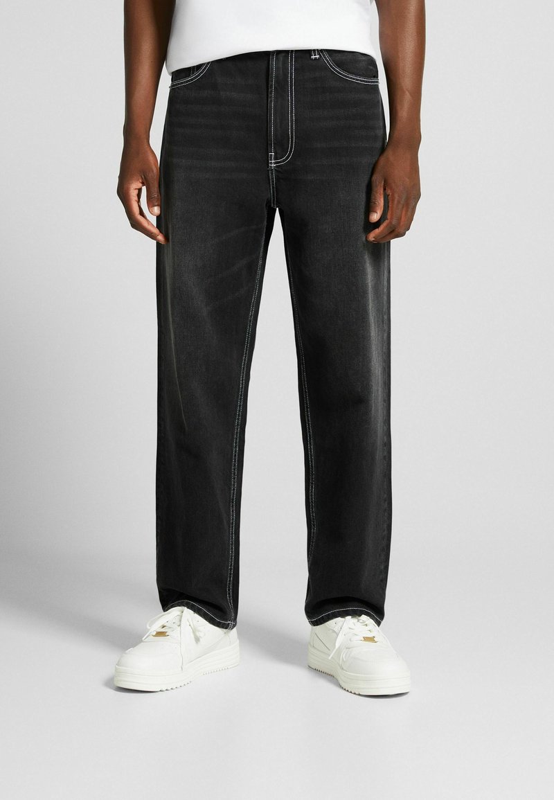 Personne portant un jean noir à jambes droites avec des coutures blanches et des baskets blanches, debout devant un fond gris.