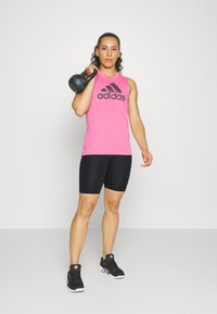 Top senza maniche rosa con logo Adidas nero, pantaloni corti neri e scarpe da ginnastica nere. Tiene in mano un kettlebell nero con dettagli blu.