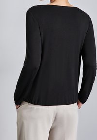 Svart långärmad topp med bred halsringning och slät textur, visad bakifrån, tillsammans med ljusbeiga byxor.