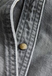 Grauer Denim-Stoff mit strukturierter Oberfläche, ausgestattet mit einem runden schwarzen Knopf mit einem marmorierten cremefarbenen Zentrum und kontrastierendem weißen Stich.