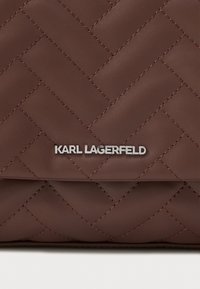 Brązowa pikowana skórzana powierzchnia z diagonalnym przeszyciem oraz srebrnym metalowym logo "KARL LAGERFELD" umieszczonym na klapie.