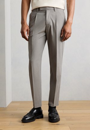 Άνδρας που φοράει ανοιχτό γκρι παντελόνι με πιέτες, μαύρες κάλτσες και μαύρα δερμάτινα μοκασίνια, στέκεται σε ξύλινο πάτωμα μπροστά σε απλή λευκή επιφάνεια.