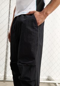 Svarta, lätta byxor med en texturerad yta, som har sidofickor och en kontrasterande grå söm längs benen.