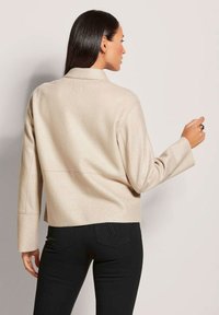 Beige, cropped jack met een gladde textuur, brede mouwen en een gelid kraag, gecombineerd met zwarte skinny jeans met een subtiele logo-detail.