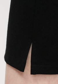 Κοντινό πλάνο μανικιού από μαύρο ύφασμα με μια μικρή πλαϊνή σχισμή, που αποκαλύπτει ελαφρύ δέρμα από κάτω και λεπτομερή ραφή κατά μήκος του τελειώματος.