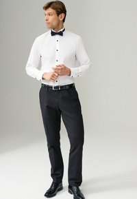 Camisa blanca de vestir con botones negros y acentos en los puños, combinada con pantalones y zapatos de vestir negros. La pajarita es de color azul marino. Diseño simple y formal.