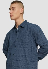 Chemise à manches longues bleu marine avec un motif géométrique, patte de boutonnage à l'avant, col et poche poitrine simple. Texture douce et coupe décontractée.