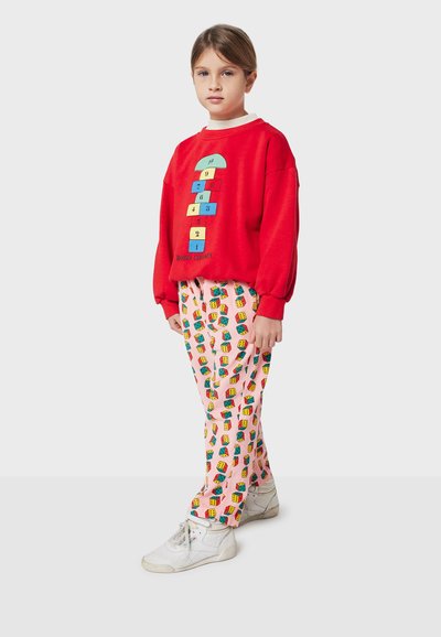 Sweat à capuche rouge avec un graphique de marelle coloré, associé à un pantalon rose présentant un motif de blocs répétitif. Des baskets blanches complètent le look.