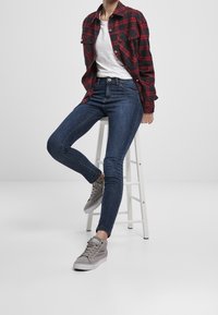 Mörkblå skinny jeans, vit t-shirt, röd och svart rutiga skjorta, grå sneakers. Modell sitter på en vit pall med en neutral bakgrund.