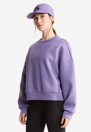 Ung kvinde iført en lavendel trøje og matchende kasket, kombineret med sorte bukser, stående mod en hvid baggrund.