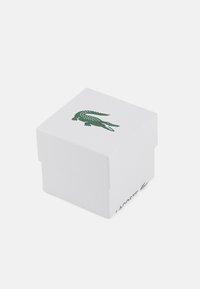 Cutie albă din carton cu o suprafață texturată, având un logo cu un crocodil verde pe capac și cuvântul "LACOSTE" pe laterale.