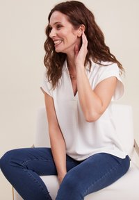 Haut blanc à manches courtes avec un col en V et une coupe décontractée, associé à un jean skinny bleu foncé. Texture lisse, sans motifs visibles ni matériel.