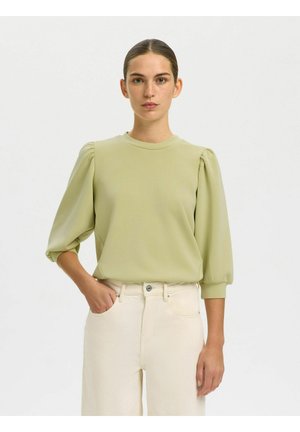 Femme aux cheveux plaqués en arrière, portant un haut vert clair à manches bouffantes et un pantalon taille haute écru, debout devant un fond uni.