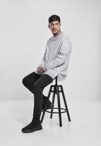 Sudadera gris de gran tamaño con cremallera lateral, combinada con pantalones negros ajustados y zapatillas negras, sentada en un taburete negro contra un fondo blanco.
