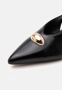 Chaussure en cuir noire à bout pointu avec un ornement doré brillant et arrondi sur le dessus près de l’avant.