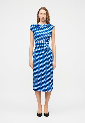 Donna in piedi che indossa un vestito blu senza maniche, lungo fino al ginocchio, con motivi ovali diagonali in nero e azzurro chiaro, abbinato a scarpe nere con punta appuntita.