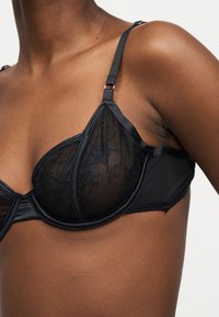 Fekete csipkés merevítős melltartó állítható pántokkal, közepesen sötét bőrön viselve, amely a felső törzset és a váll részleteit mutatja.
