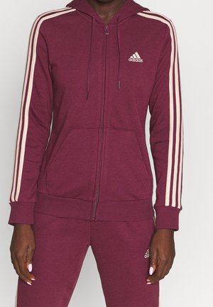 Persoon die een wijnrode Adidas-trainingspak draagt met witte strepen op de mouwen en broek, en het Adidas-logo op de borst en dij.