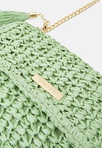 Zielona pleciona kopertówka ze złotymi okuciami, z teksturowanym wykończeniem i akcentem w postaci chwosta. Na metalowej plakietce wytłoczony napis "ANNA FIELD".