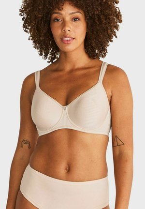 Vrouw met krullend haar die een beige beha en bijpassend ondergoed draagt, met tatoeages op beide bovenarmen, tegen een effen achtergrond.