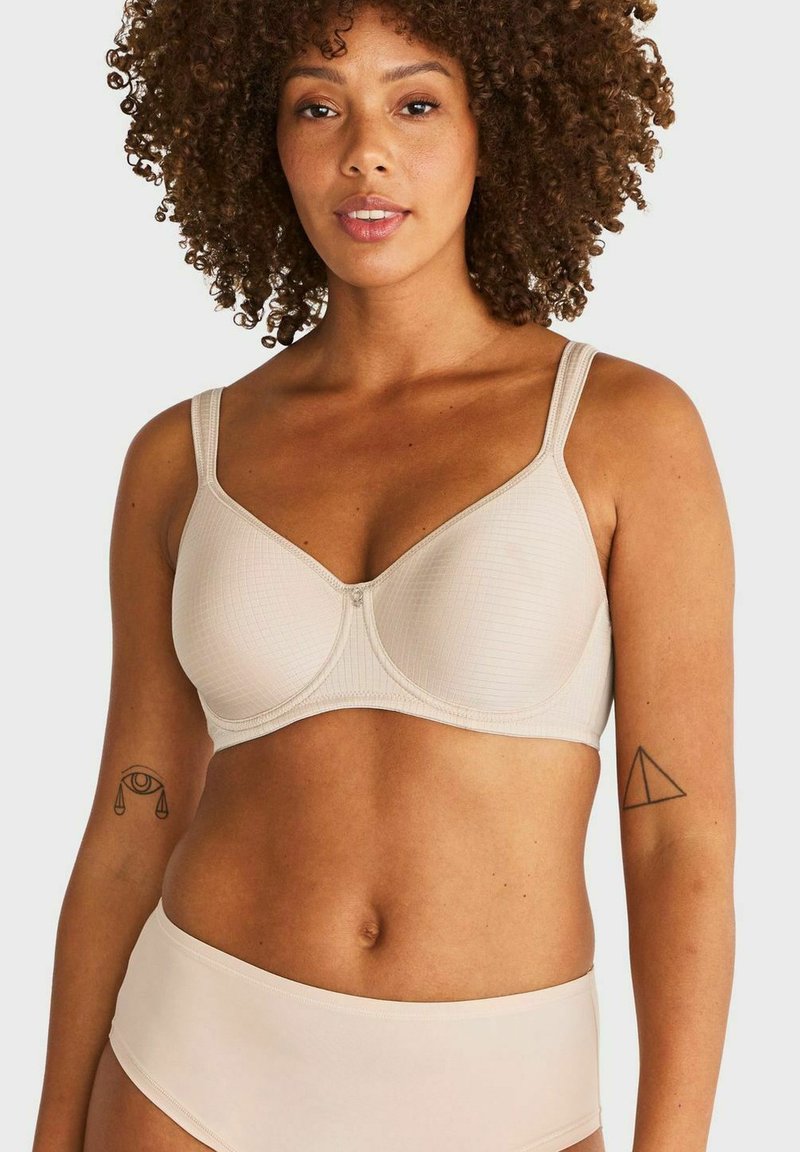 Vrouw met krullend haar die een beige beha en bijpassend ondergoed draagt, met tatoeages op beide bovenarmen, tegen een effen achtergrond.