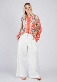 Farbenfrohe Bluse mit floralem Muster, Knopfleiste und langen Ärmeln, kombiniert mit weißen, weit geschnittenen Hosen und hellbraunen Sandalen.