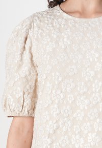 Beige bluse med pufferm, med broderte hvite blomsterdesign og en strukturert overflate. Rund hals med rynket detaljer.