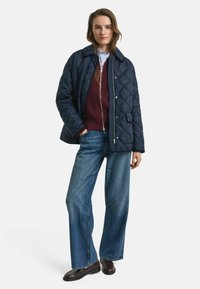 Marineblau gesteppte Jacke mit Kragen, darunter ein bordeauxroter Pullover, kombiniert mit hellblauen weiten Jeans und braunen Schuhen.